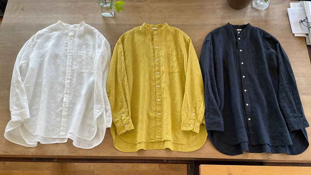 木の机に並べて置かれた 3 色のリネンシャツ。左から白、からし色、濃紺